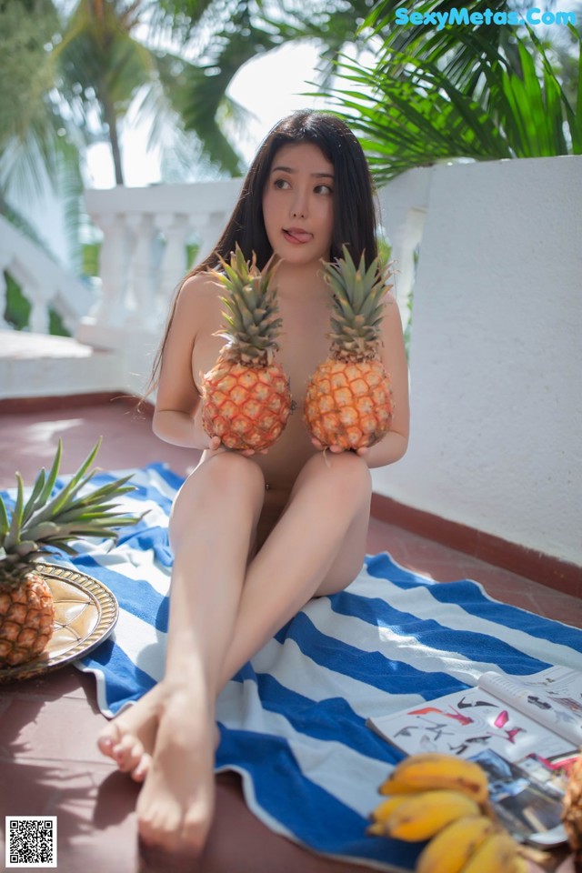 A woman sitting on a towel holding two pineapples.